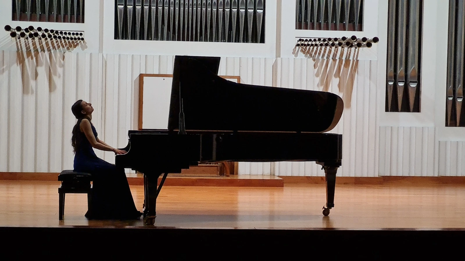Mojca Pregeljc in silhouette at a concert grand, organ pipes rising behind the stage